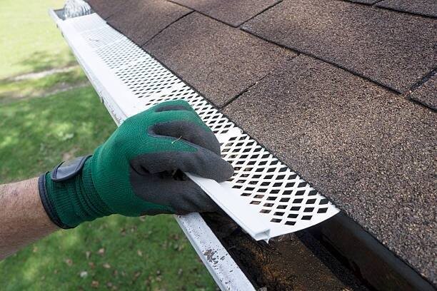 Gloved hand fitting a perforated white guard panel over a gutter at the shingle edge